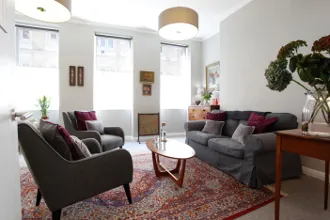 A bright psychotherapy consulting room with sofas, armchairs, soft lighting, plants, and a coffee table, creating a calm and welcoming therapy environment.