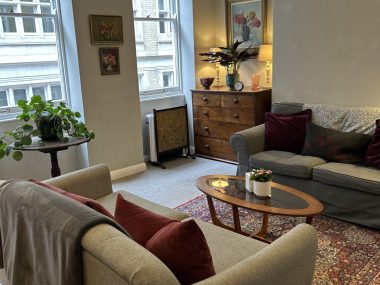 A comfortable psychotherapy consulting room with armchairs, sofas, soft lighting, plants, and a coffee table, creating a calm and welcoming therapy environment.