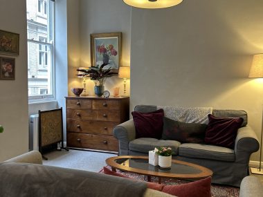 A comfortable psychotherapy consulting room with sofas, warm lighting, artwork, and soft furnishings, creating a calm and welcoming therapy environment.
